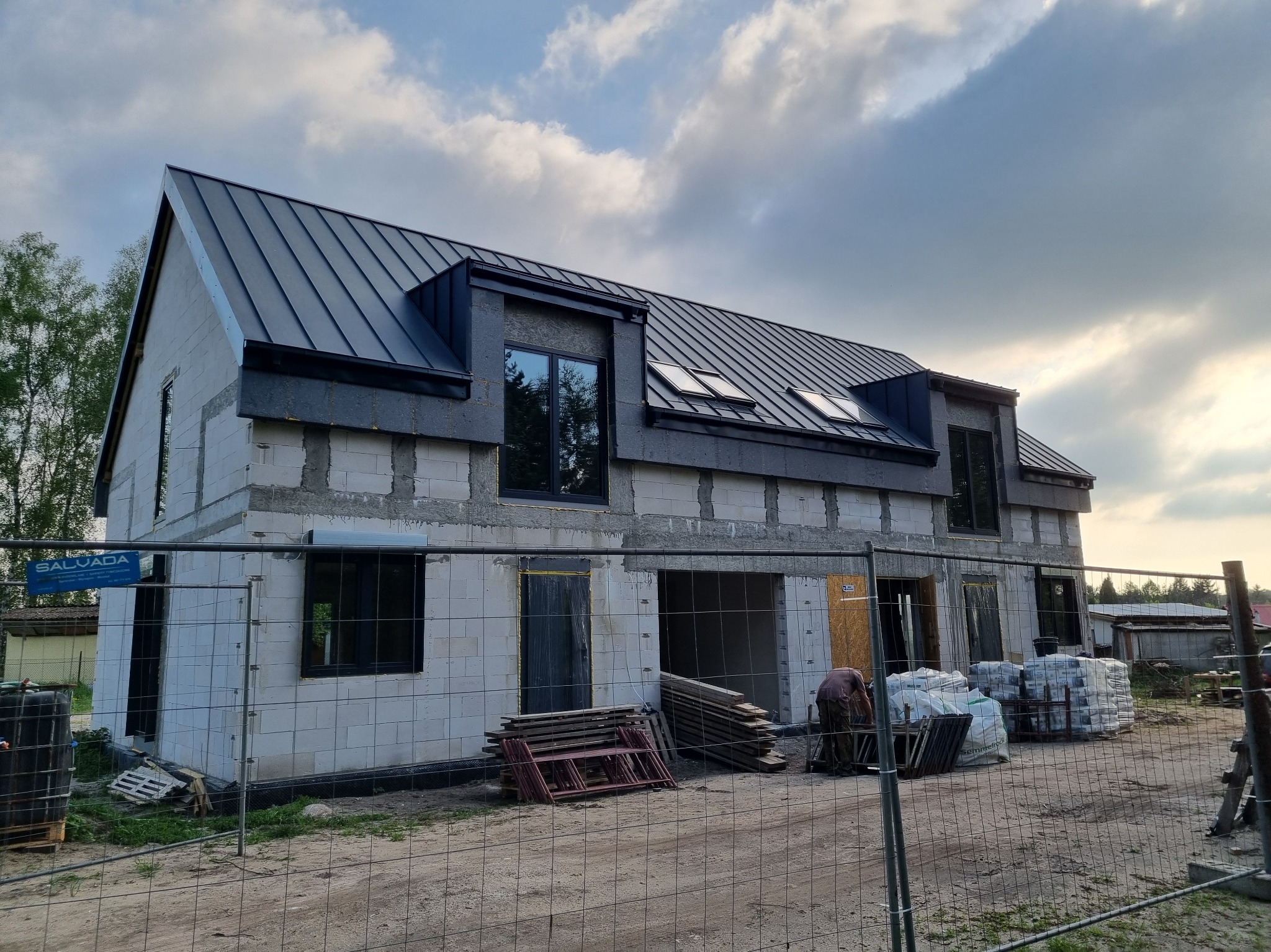 Budowa domu z białych bloczków w Goleniowie, z czarnym dachem i oknami. Widoczne materiały budowlane i ogrodzenie placu budowy. Ujęcie z dołu, pochmurne niebo.