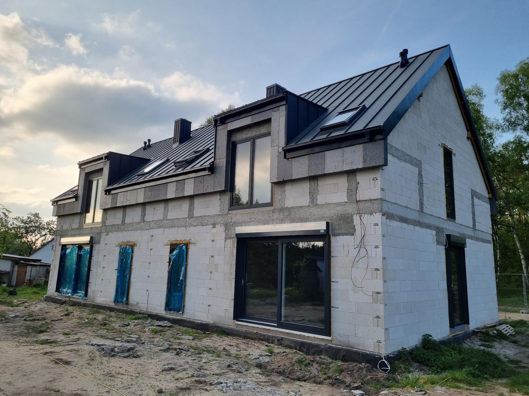 Surowy dom z betonu komórkowego z czarnym dachem i oknami. Okna zabezpieczone folią. Widoczne przewody elektryczne. Dom w trakcie budowy, otoczony ziemią.