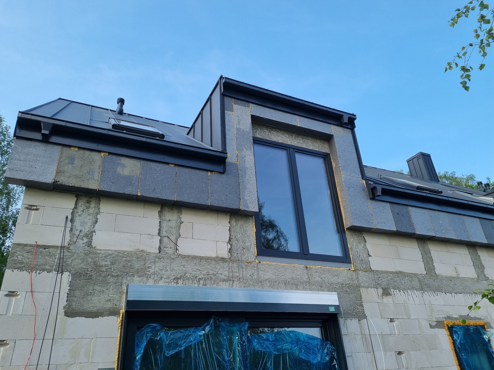 Niedokończony dom z szarej cegły z czarnym dachem i oknami. Widoczne ocieplenie i folia ochronna na oknach. Ujęcie z dołu, błękitne niebo w tle.