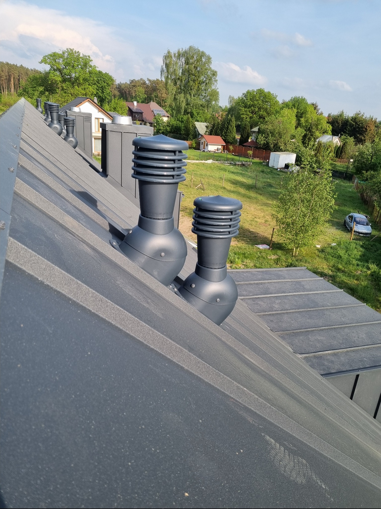 Szary dach z blachy trapezowej z zamontowanymi, nowoczesnymi kominkami wentylacyjnymi. W tle zabudowa jednorodzinna i zieleń.