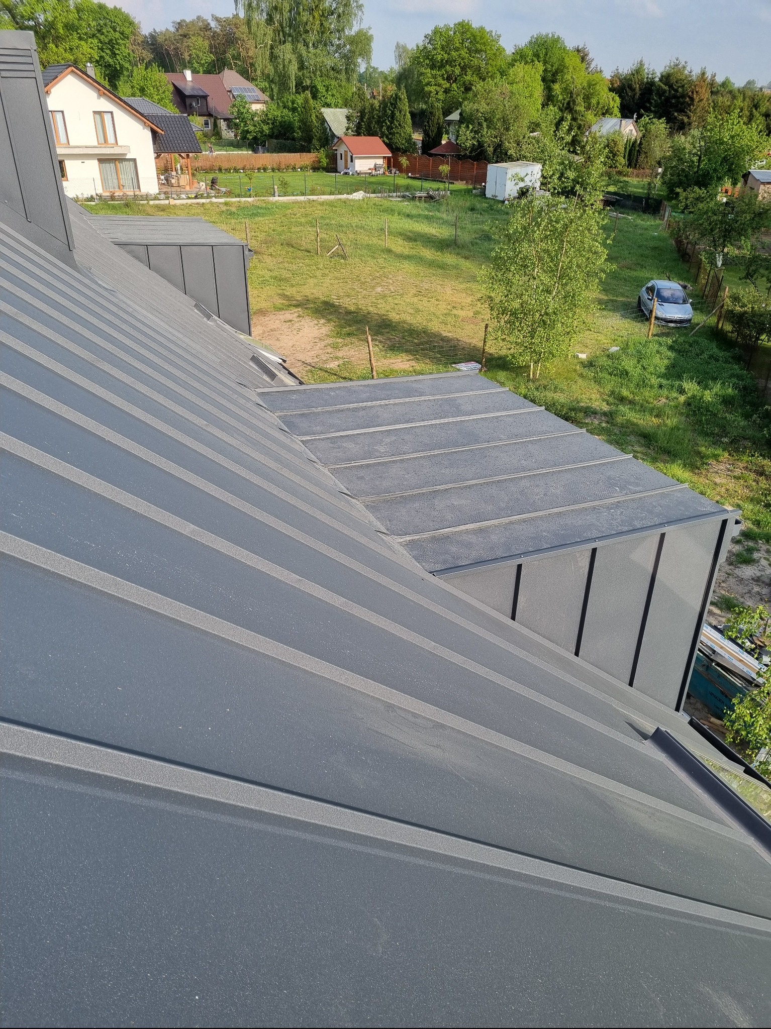 Nowoczesny, szary dach z blachy na tle zielonego ogrodu i zabudowy jednorodzinnej. Widok z góry, ukazujący detale łączeń i fakturę materiału.