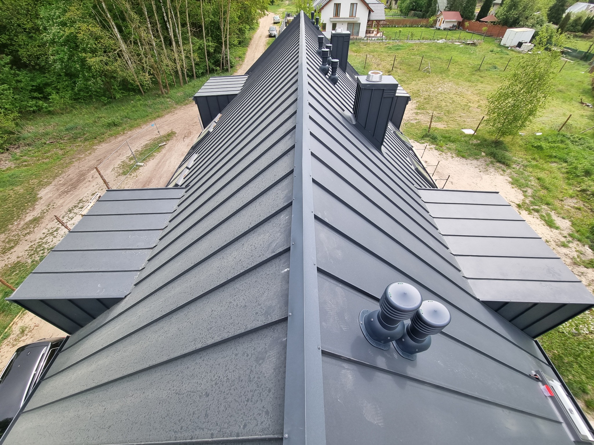Szary dach z blachy na domu jednorodzinnym, widok z góry, z kominami i wentylacją. W tle drzewa i domy. Charakterystyczne ułożenie blachy na dachu.