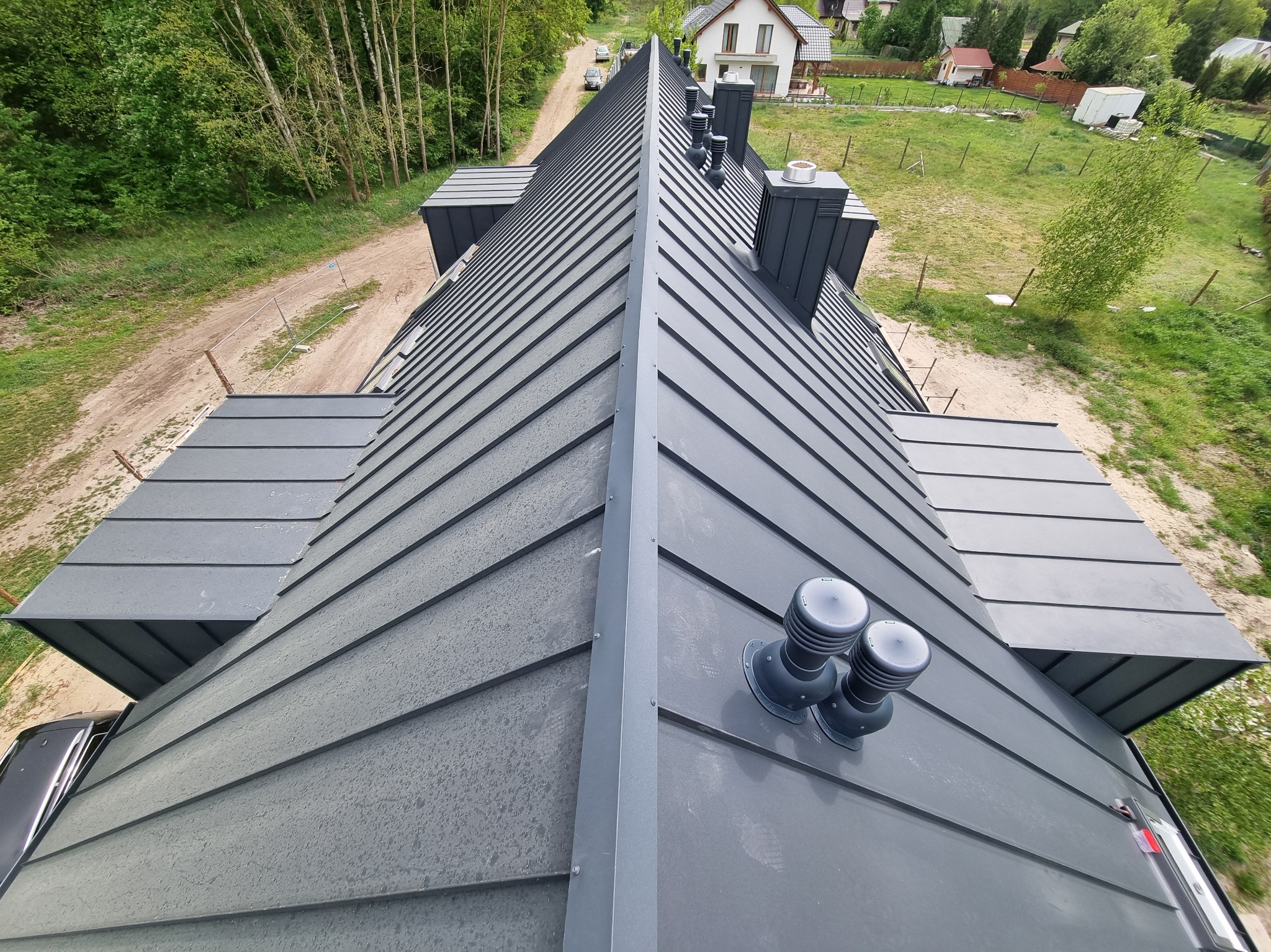 Szary dach z blachy na domu jednorodzinnym, widok z góry, z kominami i wentylacją. W tle drzewa i inne budynki. Widoczne ślady po deszczu na dachu.