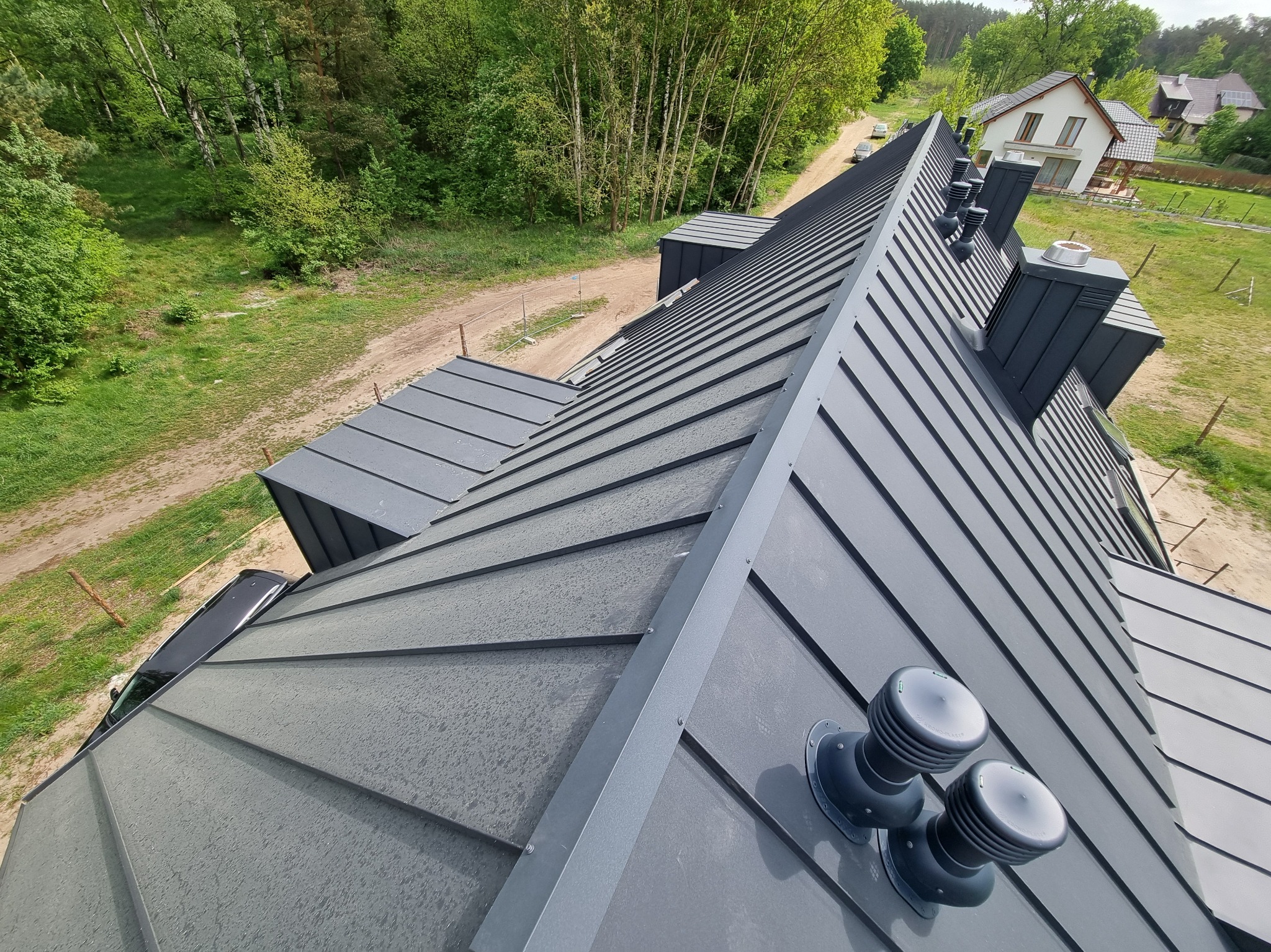 Szary dach z blachy na domu jednorodzinnym, widok z góry. Na dachu kominy i wentylatory dachowe. W tle drzewa i inny dom.