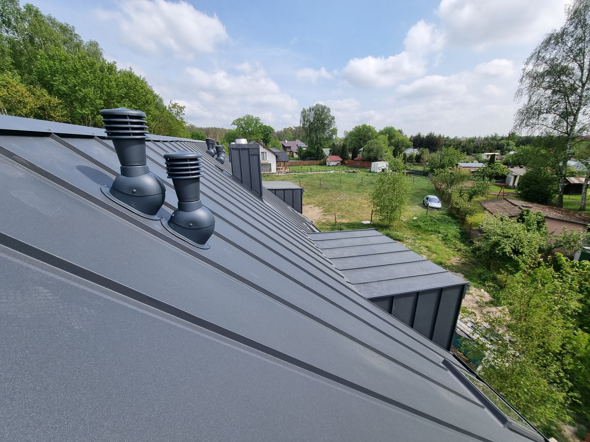 Szary dach z blachy na tle zieleni i błękitnego nieba z kominkami wentylacyjnymi. Widok z góry, ukazujący precyzję wykonania i estetykę pokrycia dachowego.