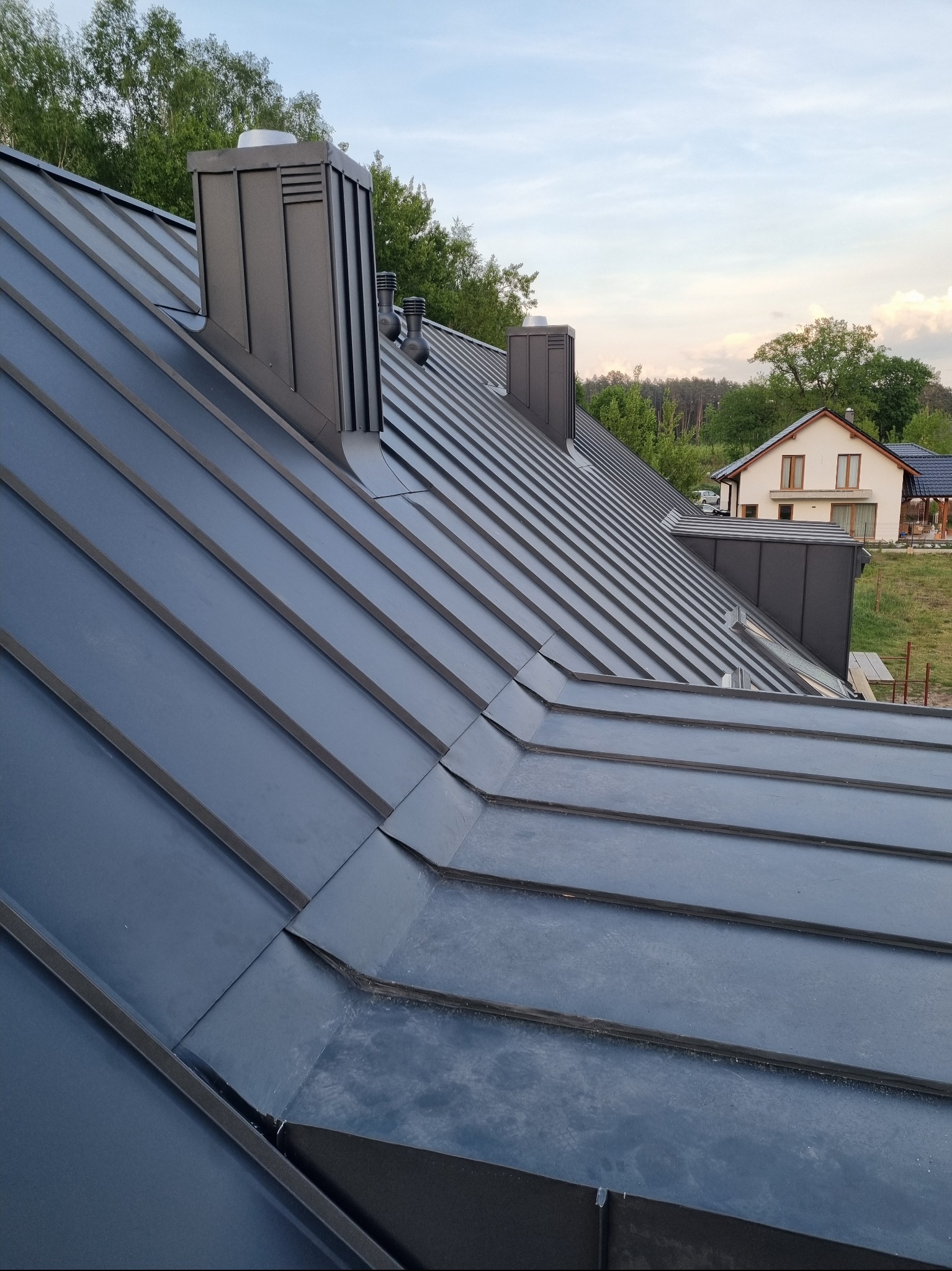 Szary, blaszany dach z kominem i lukarną na tle domu jednorodzinnego i zieleni. Widok z góry, ukazujący detale łączeń blachy i elementy wentylacyjne.
