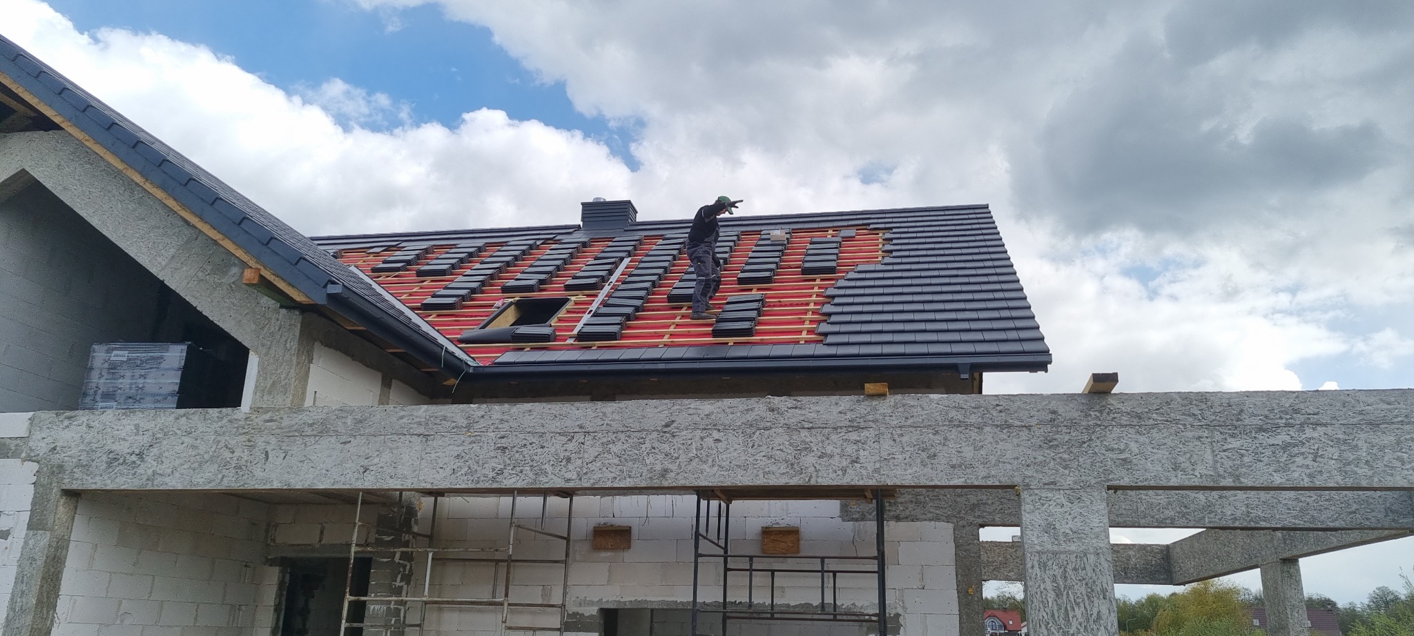 Montaż czarnej dachówki na nowym domu w trakcie budowy, widoczna więźba dachowa i dekarz pracujący na dachu, częściowo pokrytym dachówką.