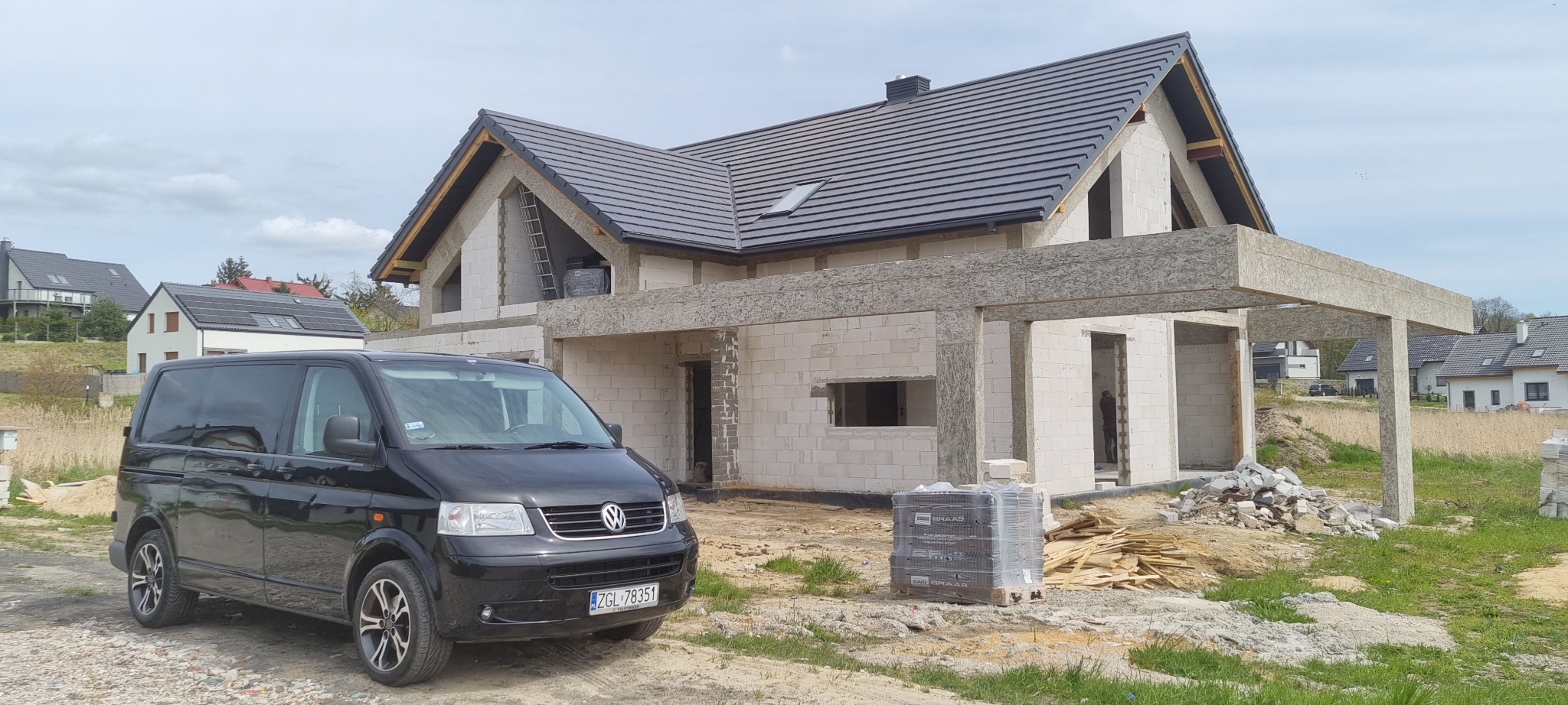 Niedokończony dom murowany z czarną dachówką, widoczny z przodu czarny van Volkswagen i paleta dachówek Braas, na drugim planie zabudowa jednorodzinna.