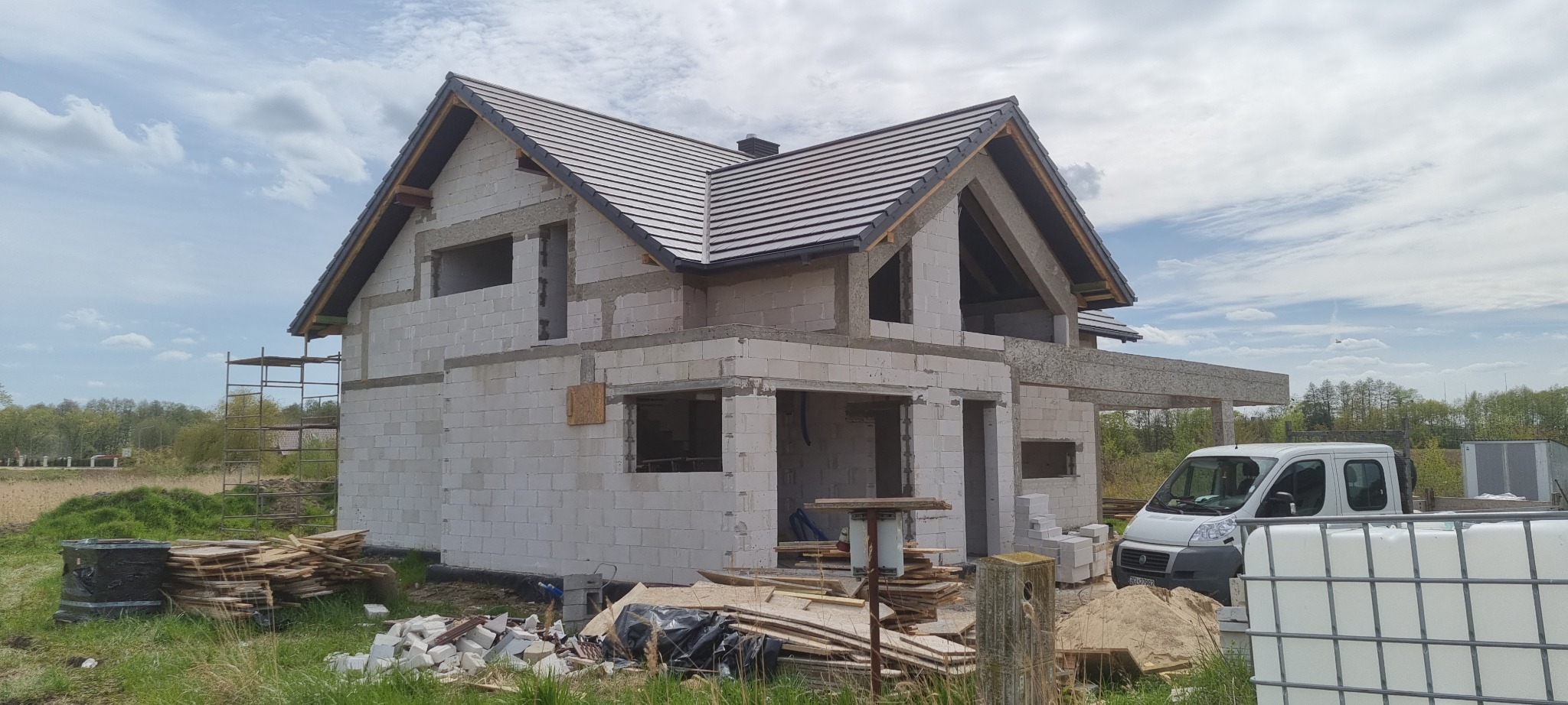 Surowy stan domu z białych bloczków, częściowo pokryty ciemną dachówką, widoczny biały bus i materiały budowlane na trawie.