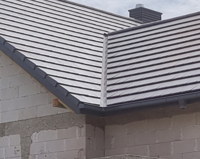 Szary dach z dachówki ceramicznej na budynku w trakcie budowy, widoczne białe bloczki i komin wentylacyjny.