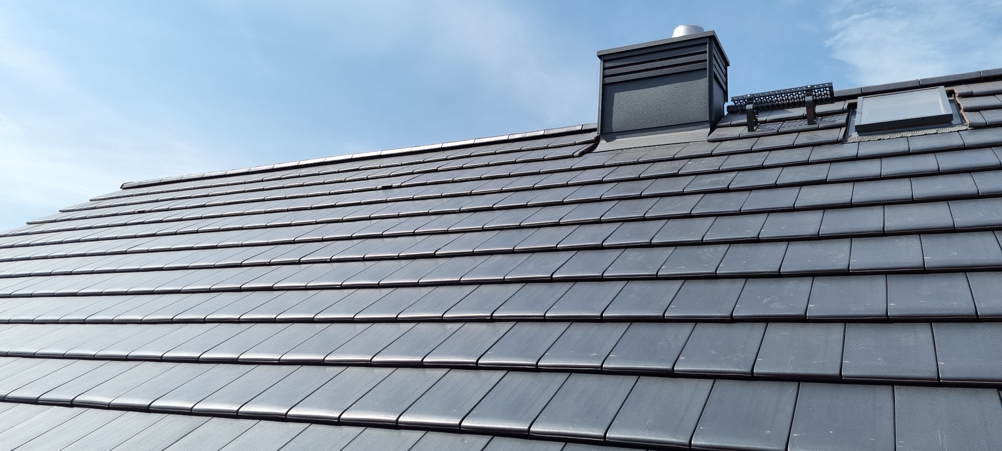 Nowy dach z ciemnoszarej dachówki ceramicznej, komin z wentylacją i okno dachowe na tle jasnego nieba.