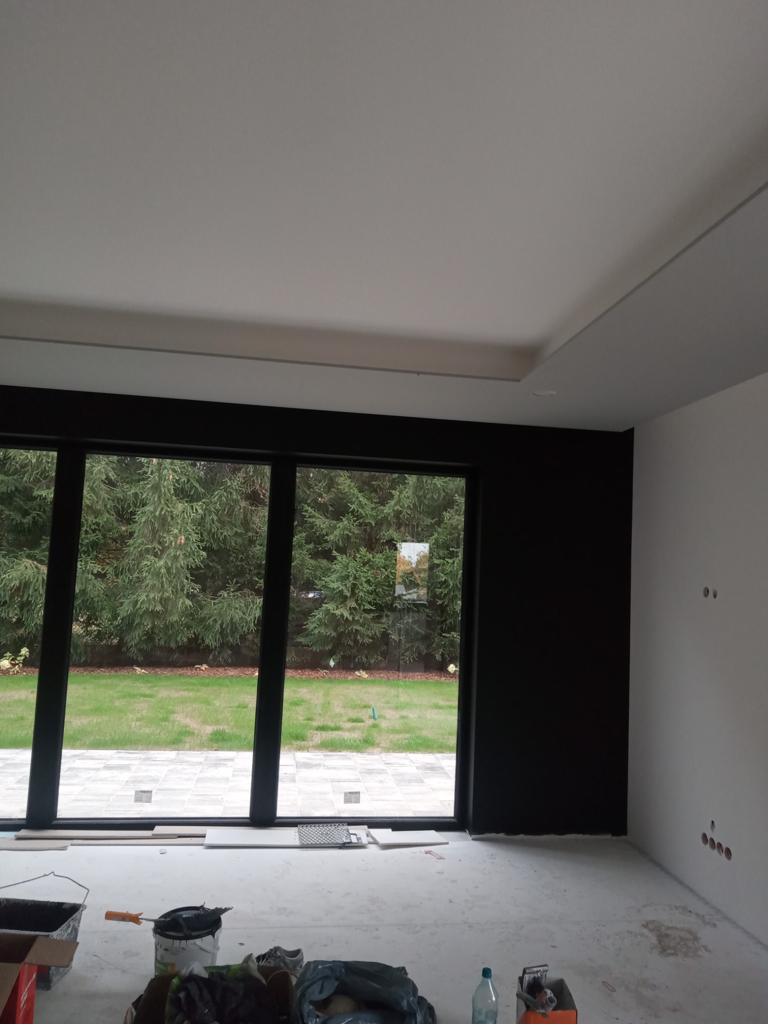 Pokój w trakcie prac wykończeniowych: świeżo pomalowana czarna ściana kontrastuje z białą ścianą i sufitem, duże okno z widokiem na ogród, widoczne narzędzia malarskie i materiały budowlane...