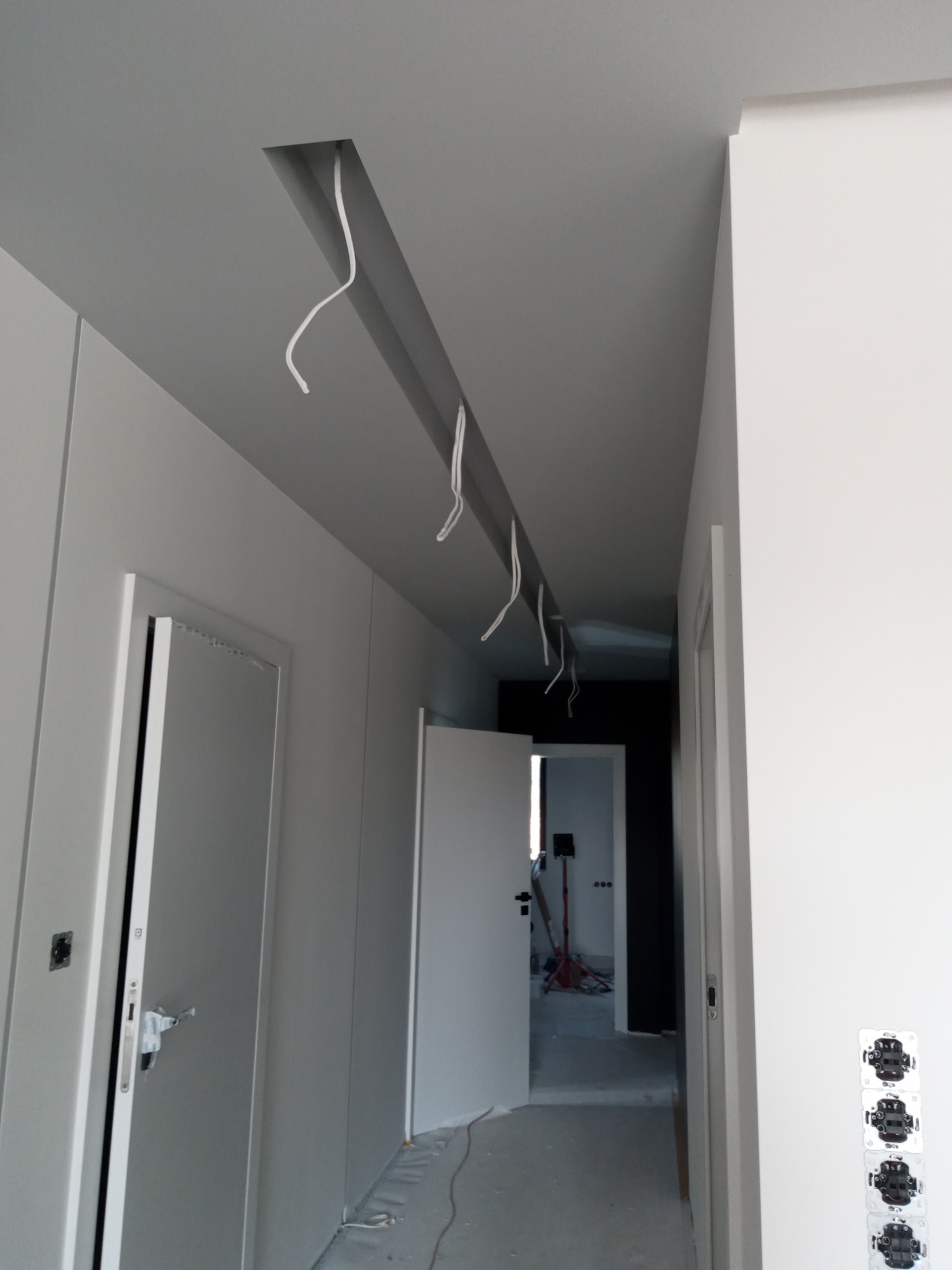 Korytarz w trakcie remontu: widoczne przewody elektryczne wystające z podwieszanego sufitu, ściany przygotowane do wykończenia, widoczne puszki elektryczne na ścianie, drzwi wewnętrzne i narzędzia...