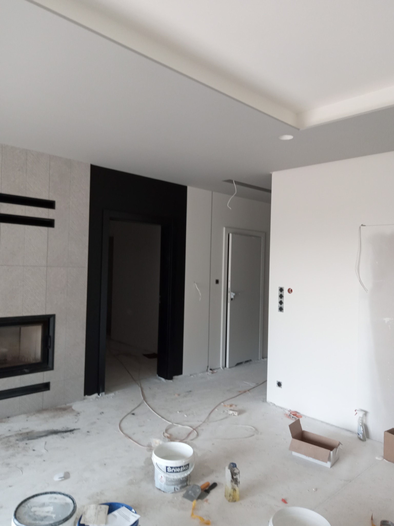 Wnętrze w trakcie remontu: świeżo pomalowane ściany w odcieniach bieli, widoczne puszki z farbą Śnieżka na podłodze, przygotowane do dalszych prac wykończeniowych, z czarnymi akcentami obramowań...