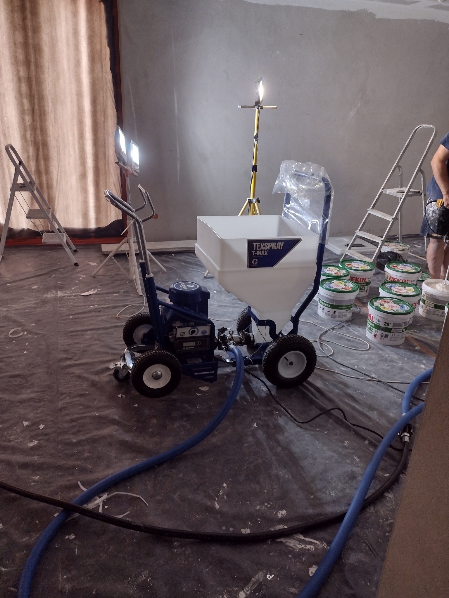 Agregat malarski TexSpray T-MAX na folii ochronnej, obok wiadra z gładzią, drabiny i lampy oświetleniowej w trakcie prac wykończeniowych wnętrza.