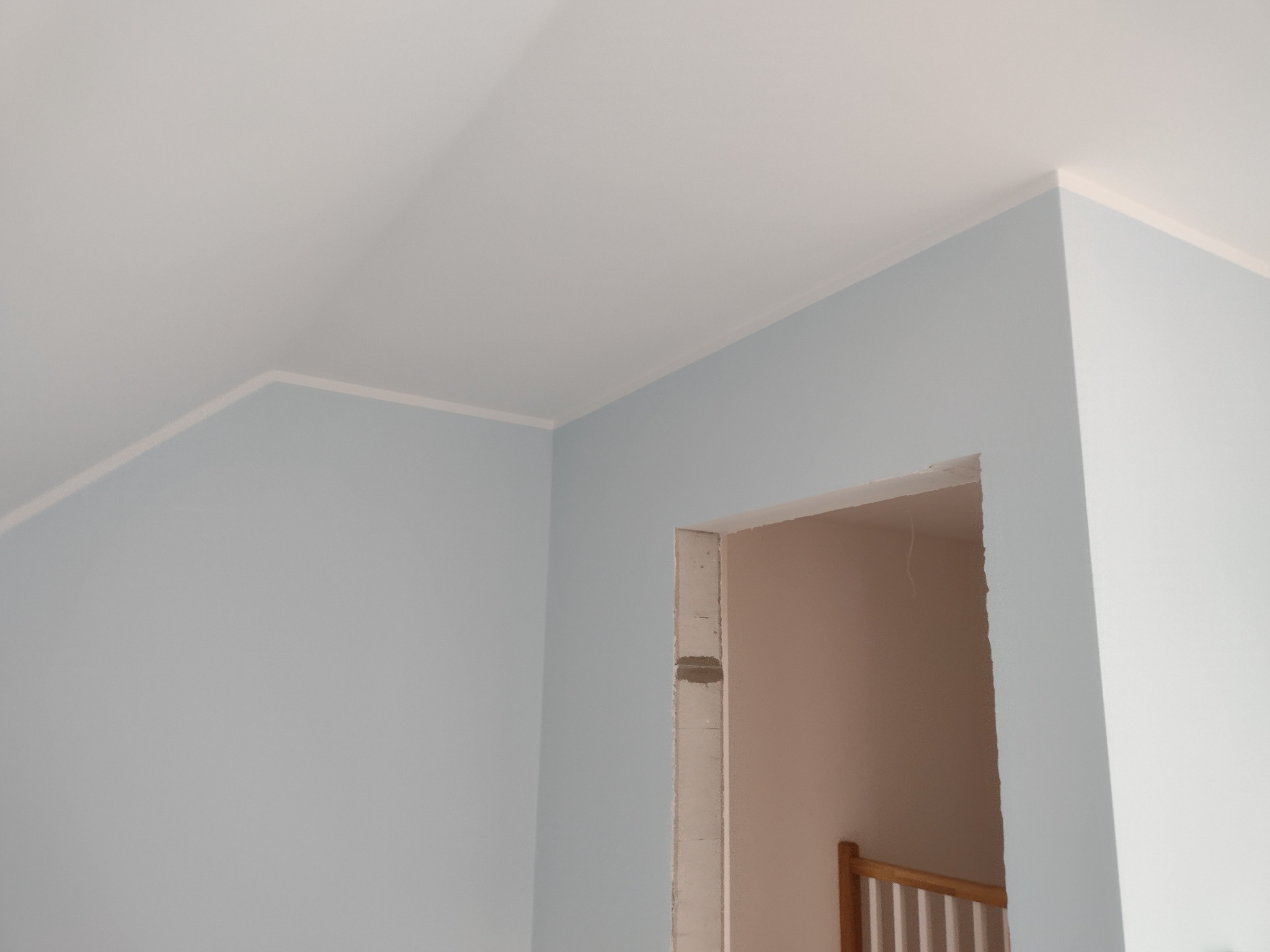 Świeżo pomalowane na jasnoniebiesko ściany w rogu pokoju z białą listwą przypodłogową i ościeżnicą drzwiową w stanie surowym, widoczna klatka schodowa w tle.