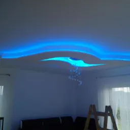 Nowoczesny sufit podwieszany z dekoracyjnym oświetleniem LED w kolorze niebieskim i elementem dekoracyjnym przypominającym krople wody, widoczna drabina malarska i fragment sofy.