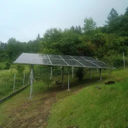 Rząd paneli słonecznych na metalowych słupach, ustawiony na wzgórzu z widokiem na zalesione zbocze w pochmurny dzień.