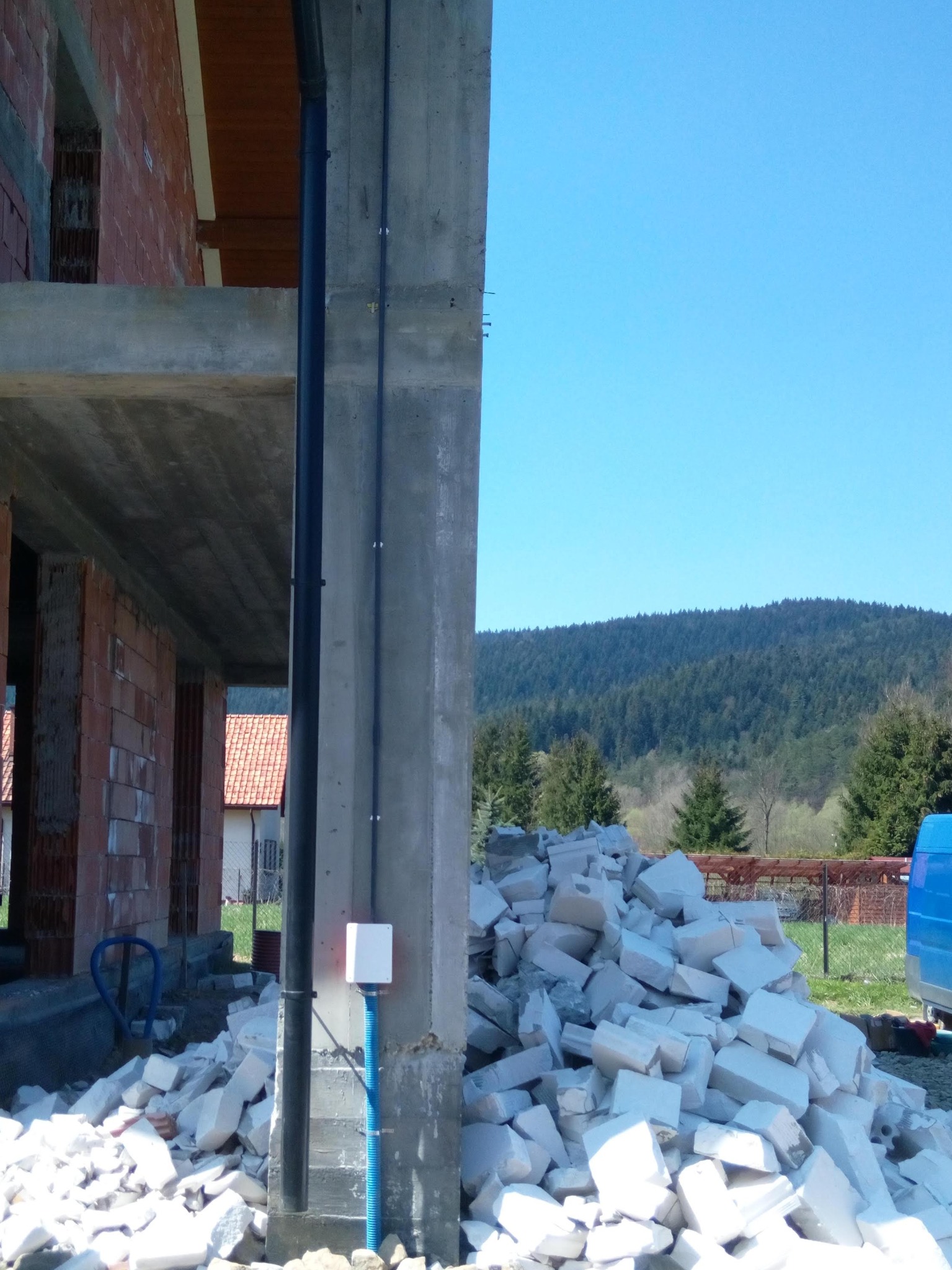 Instalacja elektryczna na surowym betonie budynku w trakcie budowy, widoczne kable w peszlu i puszka elektryczna, w tle góra gruzu i zalesione wzgórze.