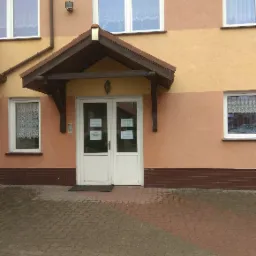 Wejście do budynku z białymi drzwiami pod zadaszeniem, na drzwiach widoczne ogłoszenia, elewacja w odcieniach beżu i brązu, przed wejściem wybrukowany chodnik.