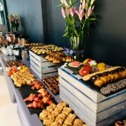 St&oacute;ł cateringowy z r&oacute;żnorodnymi przekąskami: mini kanapki z szynką parmeńską, ciastka z kruszonką, rogaliki, ciasteczka i owoce, dekorowany liliami w szklanym wazonie na tle ciemnoszarej ściany.