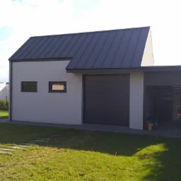 Nowoczesny budynek gospodarczy z garażem i zadaszeniem, wykończony w odcieniach szarości, z dwoma małymi oknami i metalowym dachem, otoczony trawnikiem i ścieżką z płyt.