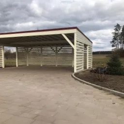 Drewniana wiata garażowa w kolorze kremowym z czerwonym dachem, widok z zewnątrz na brukowany podjazd i okoliczny krajobraz polny pod pochmurnym niebem.