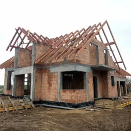 Surowy stan domu z czerwonej cegły z widoczną konstrukcją drewnianą dachu w trakcie budowy, z deskami szalunkowymi w tle, w gminie Starogard Gdański.