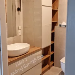 Nowoczesna łazienka z białą umywalką nablatową, drewnianym blatem i szafką z otwartymi p&oacute;łkami, szare płytki na ścianach, widoczny fragment toalety.