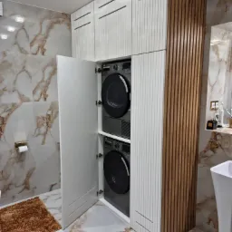 Wnęka w łazience z pralką i suszarką w zabudowie z białymi frontami i drewnianymi lamelami po boku, marmurowe płytki na ścianach i podłodze, otwarte drzwi do wnęki ukazują sprzęt.