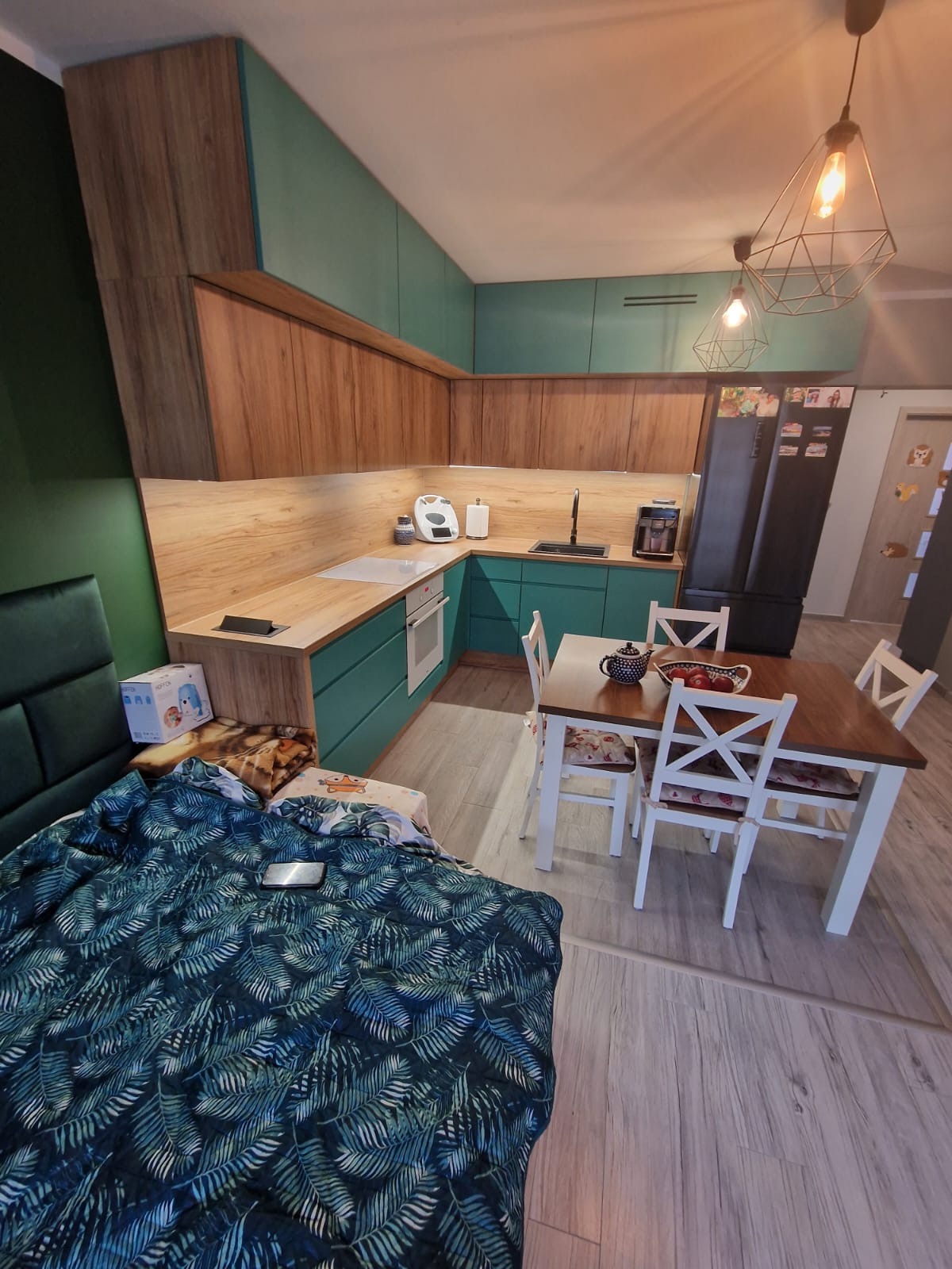 Nowoczesna kuchnia z drewnianymi i turkusowymi szafkami, oświetlona geometrycznymi lampami, widoczna z perspektywy łóżka z zielonym wzorem liści na pościeli.