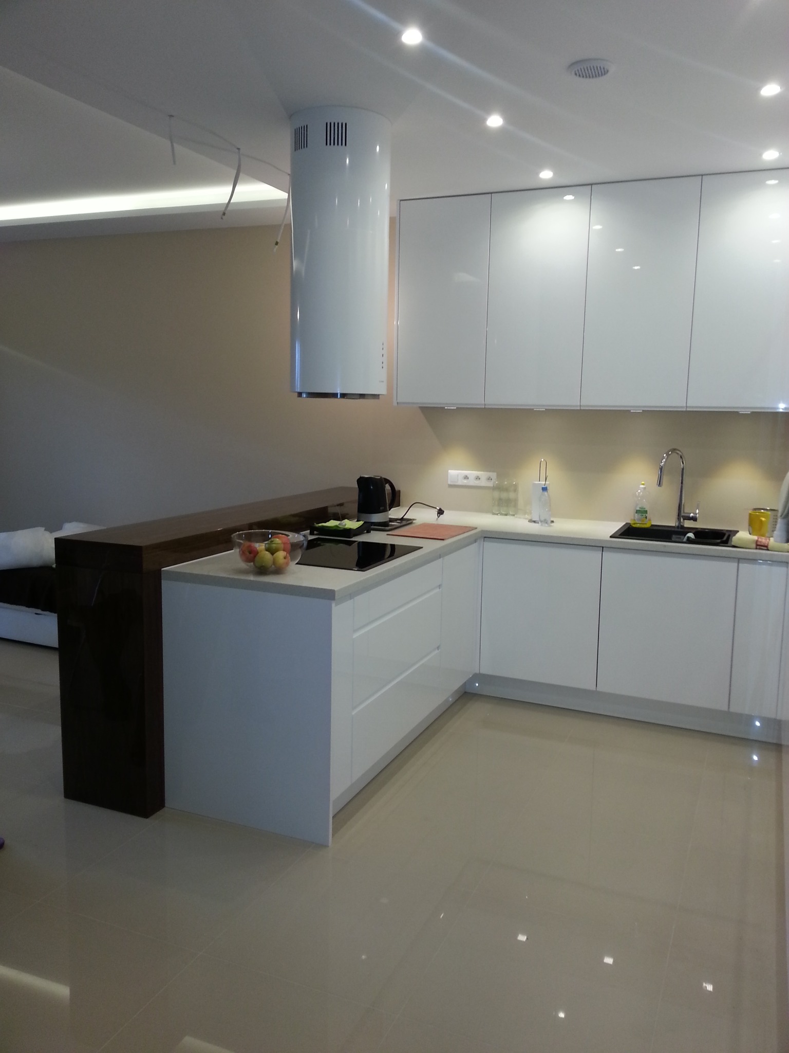 Nowoczesna kuchnia z białymi, lakierowanymi frontami, cylindrycznym okapem i blatem z misą owoców, widok na salon z sofą.