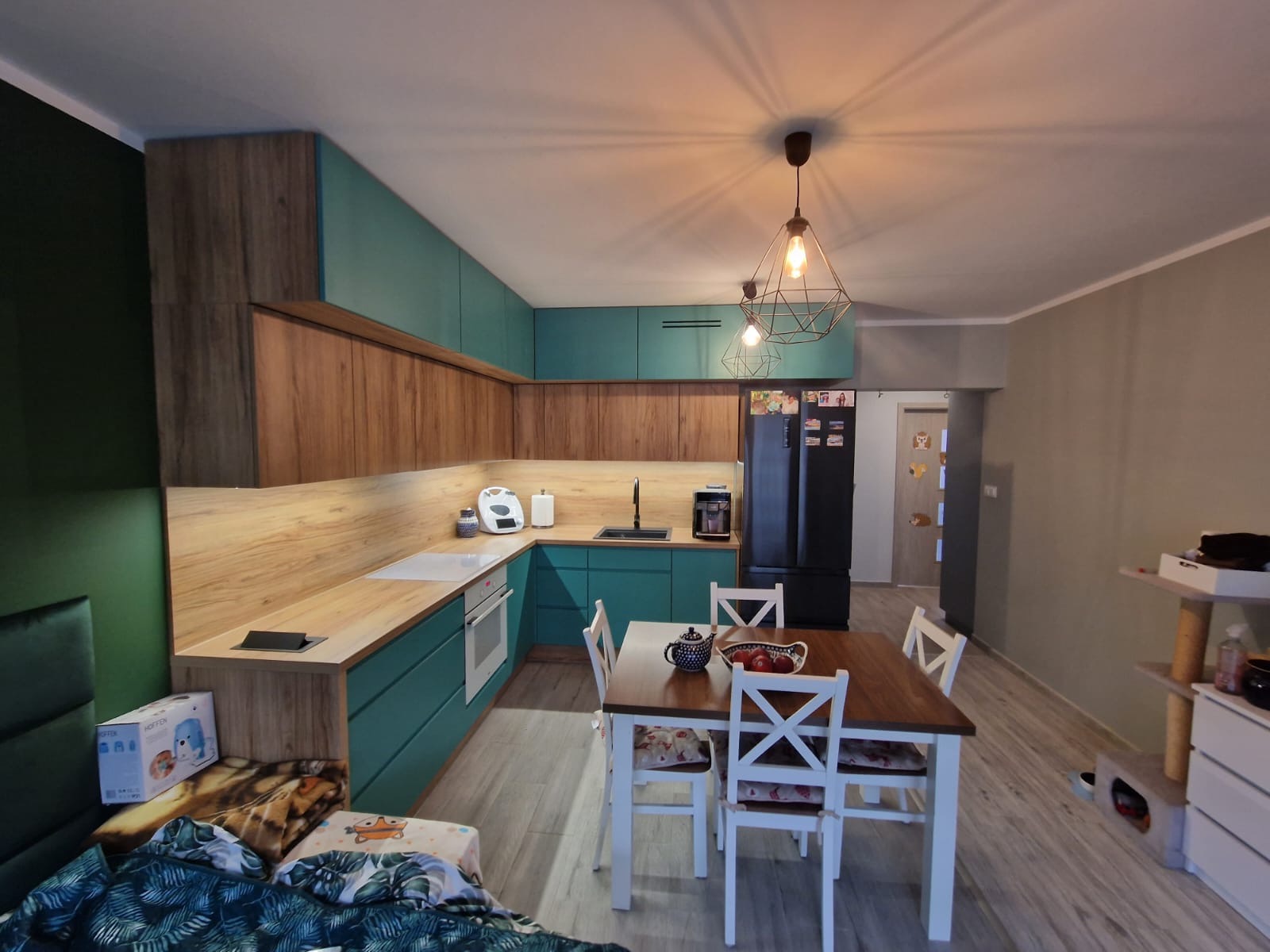 Nowoczesna kuchnia w odcieniach drewna i turkusu z drewnianym blatem, oświetlona geometryczną lampą wiszącą, z widocznym stołem jadalnym i czarną lodówką.