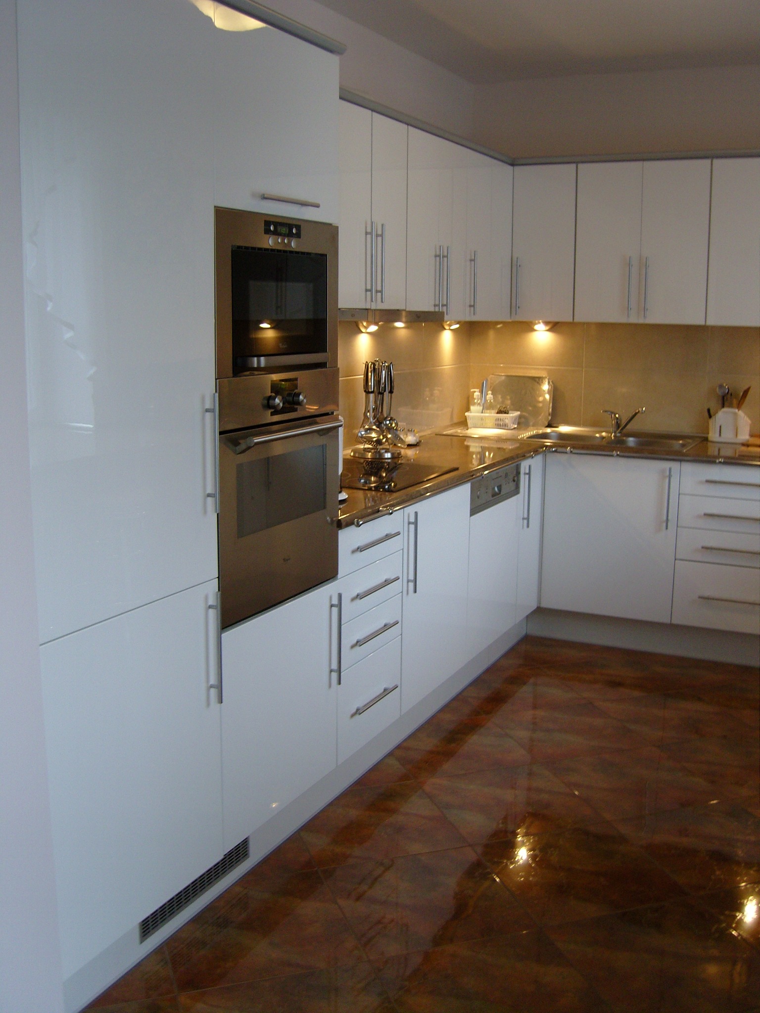 Błyszcząca, biała zabudowa kuchenna z wbudowanym piekarnikiem i mikrofalówką, oświetlona listwą LED pod szafkami, z refleksami na brązowej, lakierowanej podłodze.