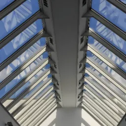 Przeszklony dach hali z widocznym niebem i chmurami, wsparty na konstrukcji stalowej z symetrycznym układem belek i mocowań.