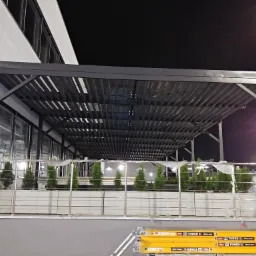 Zadaszenie tarasu z metalową konstrukcją i roślinami w donicach, widoczne rusztowanie w tle, oświetlenie nocne.