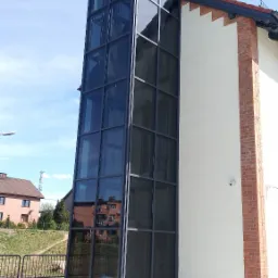 Zewnętrzna przeszklona winda osobowa, przylegająca do ściany budynku z ceglanym narożnikiem, widok z dołu na tle błękitnego nieba.