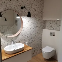 Nowoczesna łazienka z umywalką nablatową na drewnianym blacie, okrągłym lustrem, lampami ściennymi i podwieszaną toaletą, ściany wykończone płytkami imitującymi lastryko.