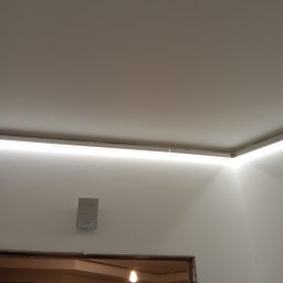 RTA Elektrosystem Radosław Kwiatkowski - Ukryte oświetlenie LED zamontowane pod sufitem w narożniku pomieszczenia, widoczny fragment ściany z zamontowanym głośnikiem i fragmentem surowej ramy drzwiowej z widoczną żarówką.