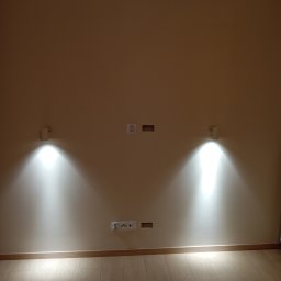 RTA Elektrosystem Radosław Kwiatkowski - Ściana z zamontowanymi dwoma kinkietami emitującymi jasne światło, gniazdkami elektrycznymi i przygotowanymi otworami na kable, nad parkietem.
