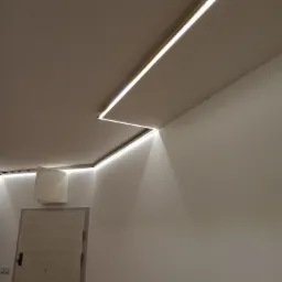 Białe ściany i sufit z zamontowanym liniowym oświetleniem LED, tworzącym geometryczny wzór wzdłuż krawędzi, widoczny fragment drzwi z zabezpieczeniem folią.