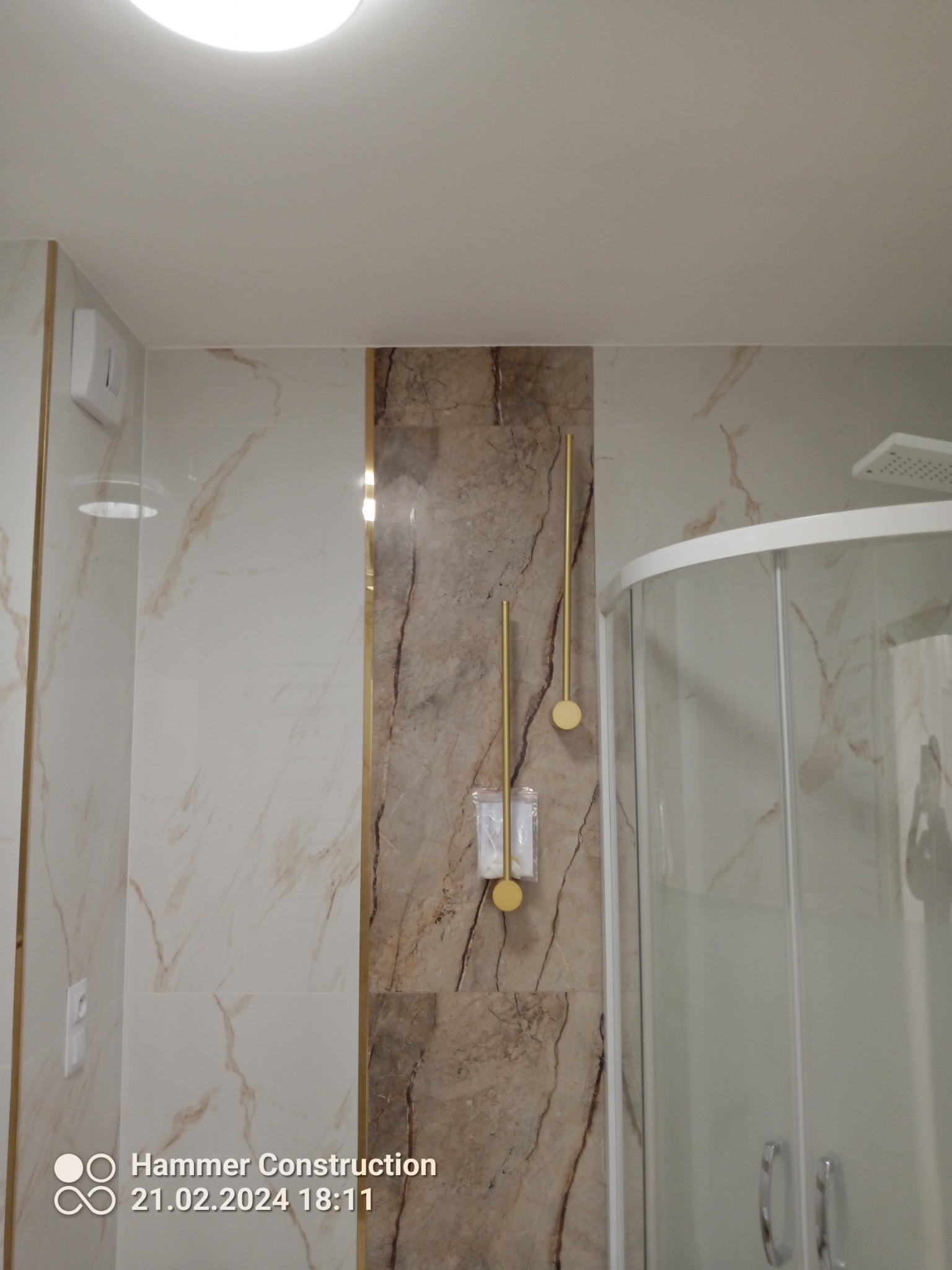 Nowoczesna łazienka z kabiną prysznicową, marmurowymi płytkami i złotymi akcentami, w tym uchwyt na papier toaletowy i włącznik światła.