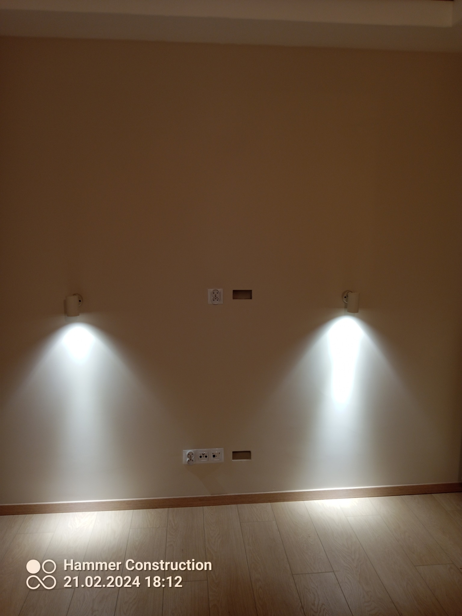 Ściana z dwoma kinkietami emitującymi jasne światło, gniazdkami elektrycznymi i pustymi otworami na przewody, świeżo położona podłoga z paneli.