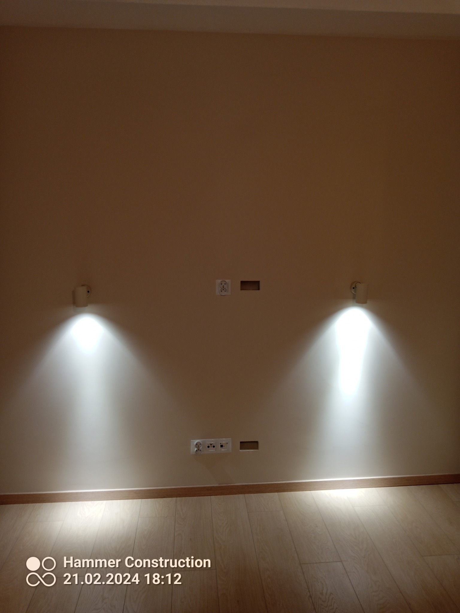 Zamontowane dwa białe kinkiety oświetlające ścianę w pokoju, widoczne gniazdka elektryczne i otwory na kable, parkiet na podłodze.