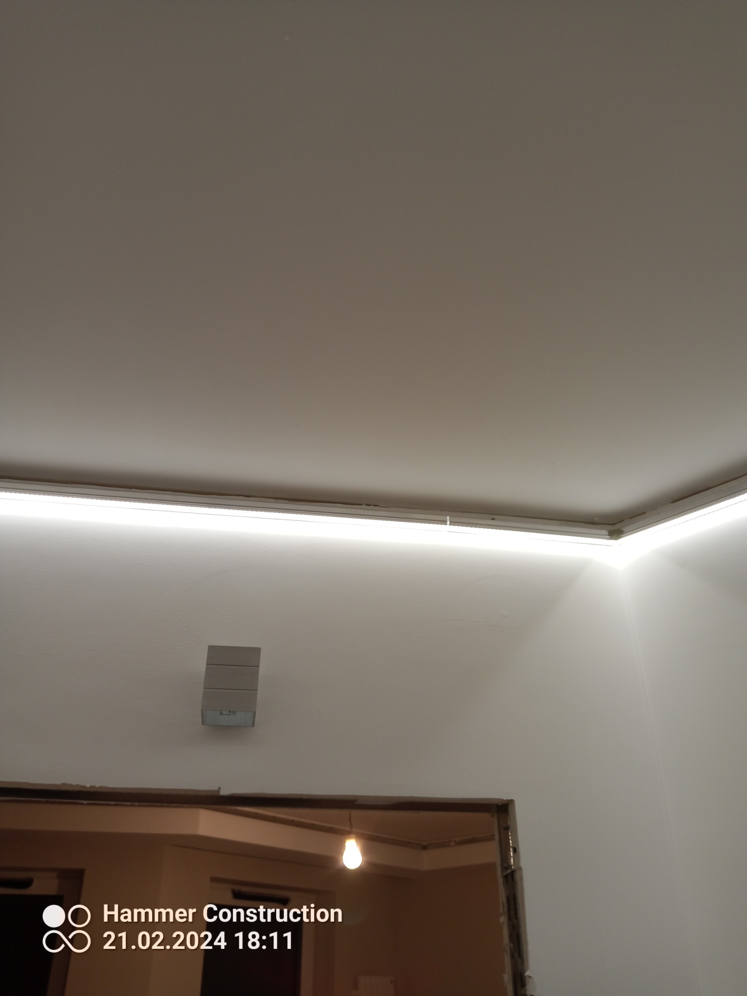 Ukryte oświetlenie LED zamontowane pod sufitem w narożniku pomieszczenia, widoczny fragment ściany z zamontowanym głośnikiem i fragmentem surowej ramy drzwiowej z widoczną żarówką.