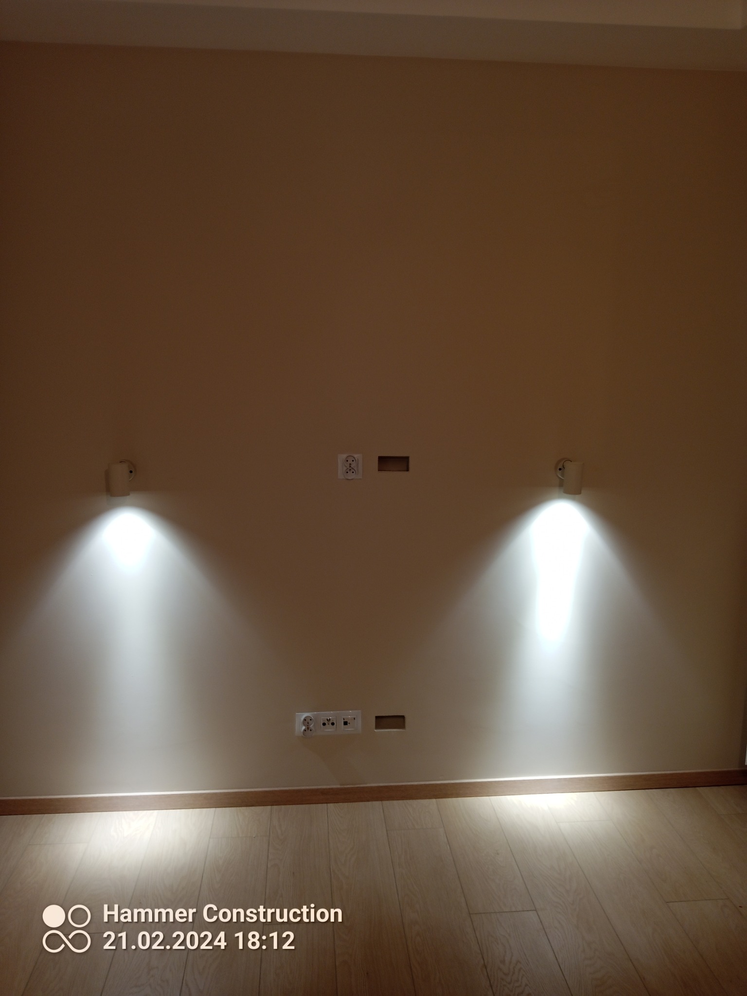 Ściana z zamontowanymi dwoma kinkietami emitującymi jasne światło, gniazdkami elektrycznymi i przygotowanymi otworami na kable, nad parkietem.