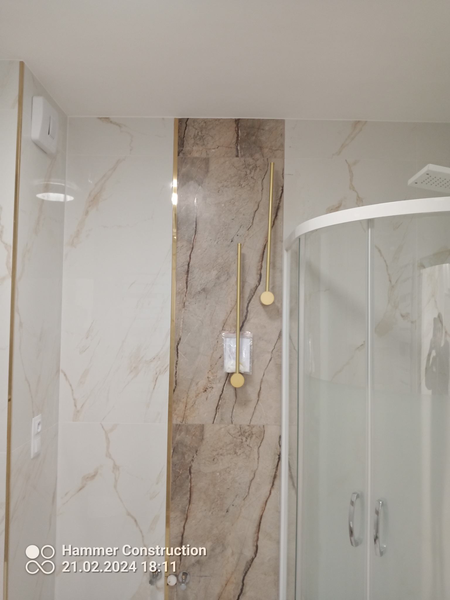 Nowoczesna łazienka z kabiną prysznicową, marmurowymi płytkami w odcieniach beżu i złota oraz złotymi akcesoriami łazienkowymi, widoczne detale wykończenia.