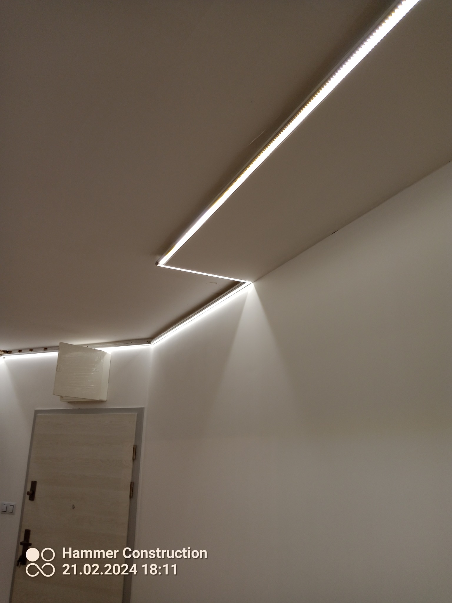 Białe ściany i sufit z zamontowanym liniowym oświetleniem LED, tworzącym geometryczny wzór wzdłuż krawędzi, widoczny fragment drzwi z zabezpieczeniem folią.