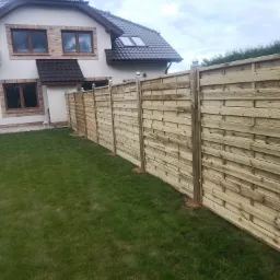 Nowoczesne ogrodzenie z jasnego drewna, w stylu poziomych paneli, z metalowymi kulami na słupkach, w Kolin, na tle domu z ceglanymi elementami.