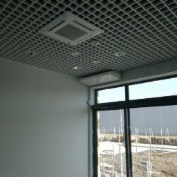 Sufit rastrowy z wbudowaną kasetą klimatyzacyjną w pomieszczeniu biurowym, widok z dołu na zainstalowaną jednostkę, widoczne elementy instalacji i okno z widokiem na teren budowy.